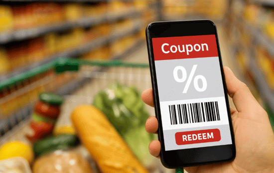 Eine Hand h&auml;lt ein Smartphone mit digitalem Coupon in einem Supermarkt, im Hintergrund ein voller Einkaufswagen mit Lebensmitteln.
