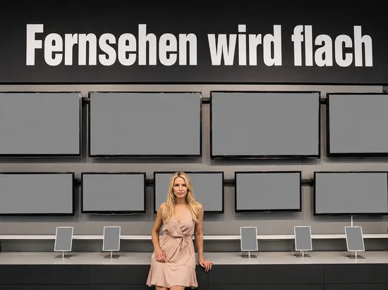 Blonde Frau im Elektronikmarkt vor einer Wand mit vielen Flachbildfernsehern, deren Bildschirme leer sind