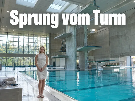Schwimmhalle mit Sprungturm und blauem Becken, eine Frau steht am Beckenrand